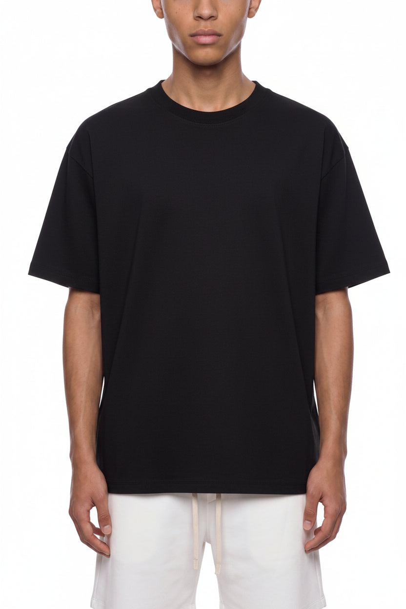 A man wearing an oversized black crewneck t-shirt and off-white pants, photographed against a solid white background.